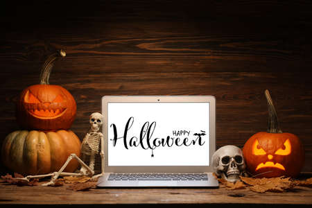 Carved pumpkins, skeleton, skull and laptop with text HAPPY HALLOWEEN on screen against wooden backgroundの写真素材