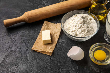 Bowl of flour, butter, chicken egg and rolling pin on dark background