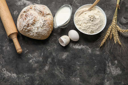 Fresh bread, bowl of flour, milk and rolling pin on dark backgroundの写真素材