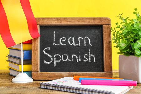 Chalkboard with text LEARN SPANISH, flag, stationery and flowerpot on table against yellow backgroundの写真素材