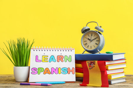 Notebook with text LEARN SPANISH, stationery, alarm clock, flowerpot and flag on table against yellow backgroundの写真素材