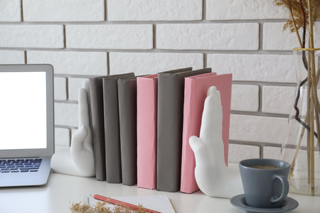Stylish holder with books, laptop, cup and vase on table near white brick wall
