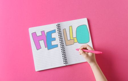Child writing word HELLO in notebook on pink backgroundの写真素材