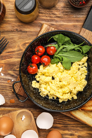 Frying pan with scrambled eggs, tomatoes and greens on wooden tableの写真素材