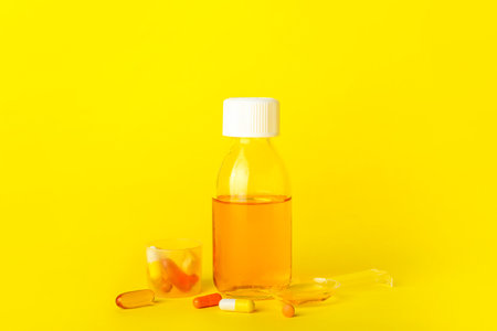 Bottle of cough syrup, cup, spoon and pills on yellow backgroundの写真素材