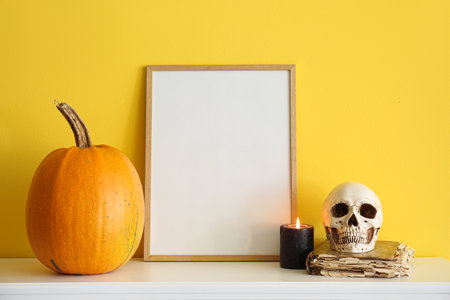 Blank frame with Halloween pumpkin, candle, skull and book on mantelpiece near yellow wallの写真素材