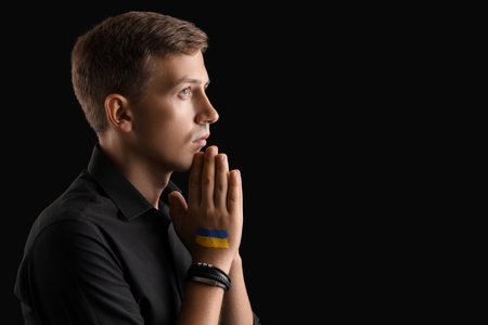 Young man with drawn flag of Ukraine praying on black backgroundの写真素材