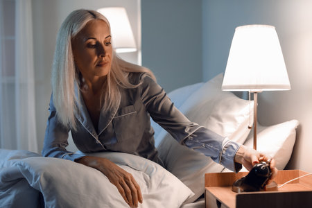 Stressed mature woman with alarm clock sitting on bed at night