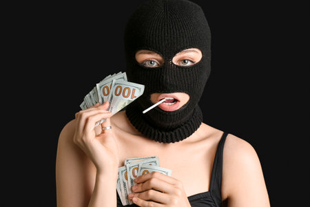 Young woman in balaclava with lollipop holding money on black backgroundの写真素材