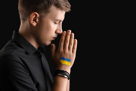 Young man with drawn flag of Ukraine praying on black backgroundの写真素材