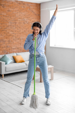 Young housewife with headphones and mop singing at home
