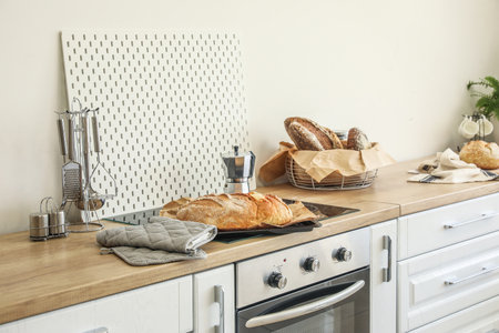 Baking sheet with fresh bread and oven mittens on stove in kitchen