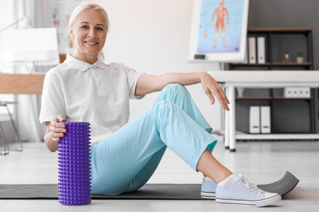 Mature physiotherapist with foam roller sitting on mat in rehabilitation centerの写真素材