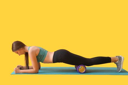 Young woman exercising with purple foam roller on yellow backgroundの写真素材