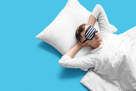 Young man sleeping mask, pillow and blanket lying on blue backgroundの写真素材
