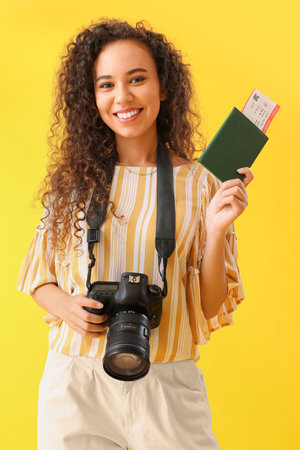 Young African-American woman with passport and photo camera on yellow background