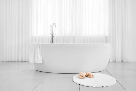 Modern bathtub and slippers on rug in light bathroomの写真素材