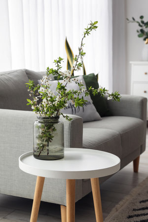 Vase with blooming tree branches on table near sofa in living room