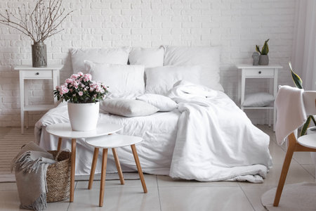 Beautiful begonia flowers and comfortable bed near white brick wall in room interiorの写真素材