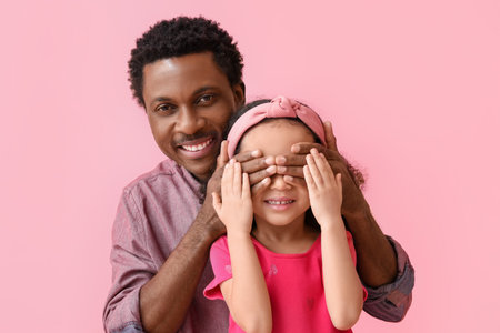 Portrait of African-American man covering eyes of his little daughter with hands on pink backgroundの写真素材