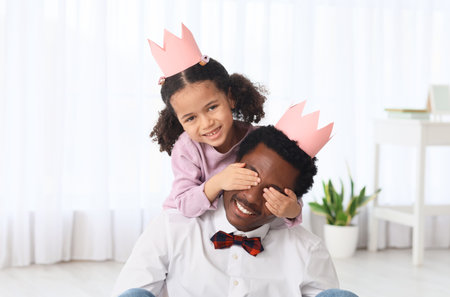 Happy African-American man and his little daughter in paper crowns playing at homeの写真素材