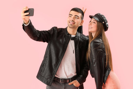 Fashionable couple in autumn clothes taking selfie on pink background