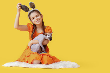 Little girl in bunny ears with Sphynx cat on yellow backgroundの写真素材