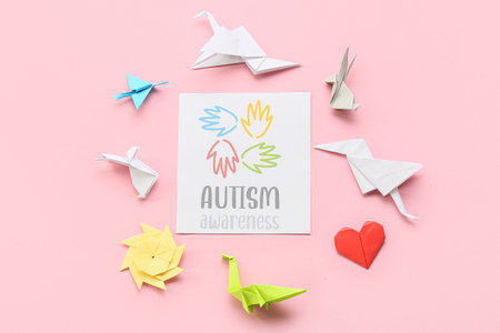 Paper sheet with text AUTISM AWARENESS, human palms and origami on pink backgroundの写真素材