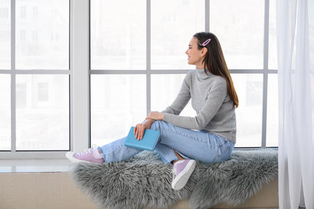 Pretty young woman with book sitting on window sill