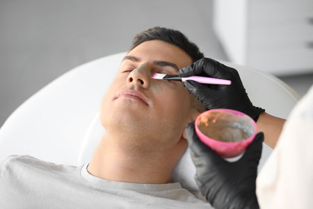 Cosmetologist applying clay mask onto young man's face in salon, closeupの写真素材