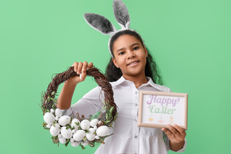 Cute African-American girl with bunny ears, greeting card and Easter wreath on color backgroundの写真素材