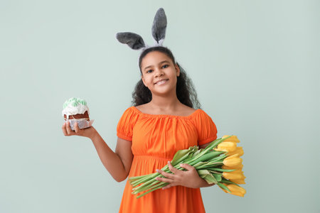 Cute African-American girl with bunny ears, flowers and Easter cake on color backgroundの写真素材