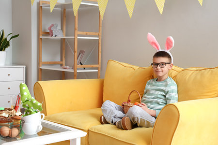 Cute little boy with bunny ears and Easter basket sitting on sofa at homeの写真素材