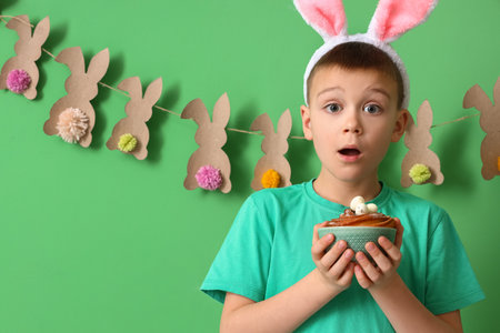 Surprised little boy with bunny ears and Easter cake on green background