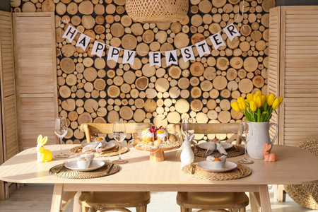 Dining table with stylish setting and garland with text HAPPY EASTER on wooden wall