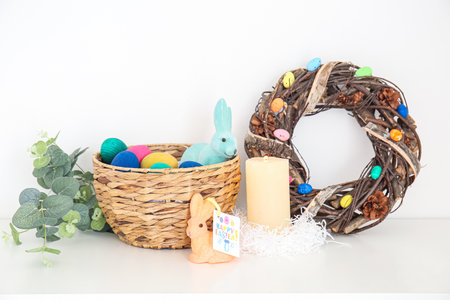 Basket with Easter eggs, rabbits, candle, eucalyptus and wreath on shelf near light wall