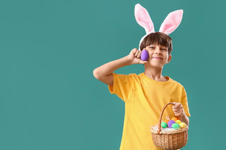 Cute little boy with bunny ears and Easter eggs in basket on color background