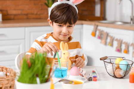 Cute little boy with figurine of bunny during painting of Easter eggs in kitchen at homeの写真素材