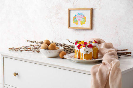 Easter cakes, bowl with eggs and pussy willow branches on chest of drawers near light wallの写真素材