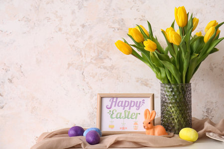 Vase with tulips, Easter eggs and photo frame on table near light wall
