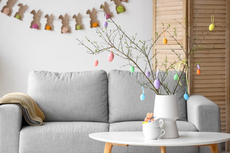Tree branches decorated with Easter eggs in vase on table near sofa