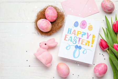 Beautiful Easter composition with greeting card, painted eggs, bunny and flowers on light wooden backgroundの写真素材