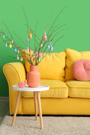 Tree branches with Easter eggs in vase on table and sofa near color wall in room