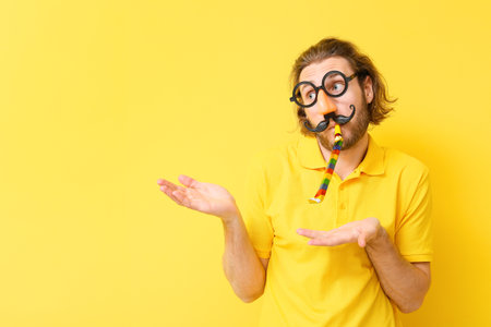 Funny young man in disguise and with party whistle showing something on yellow background. April fools' day celebrationの写真素材