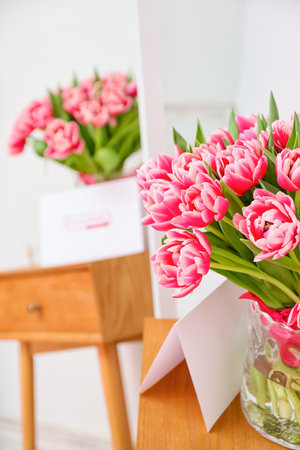 Vase with beautiful tulips and greeting card on wooden table near light wall. International Women's Day celebrationの写真素材