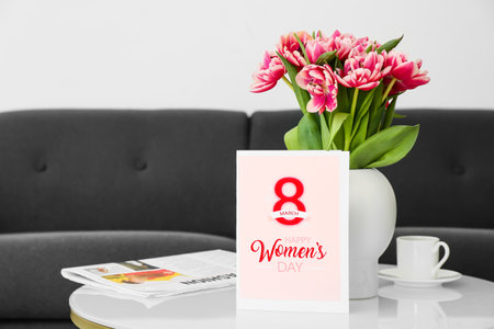 Greeting card with text HAPPY WOMEN'S DAY, vase with tulips, cup of coffee and newspaper on table in roomの写真素材
