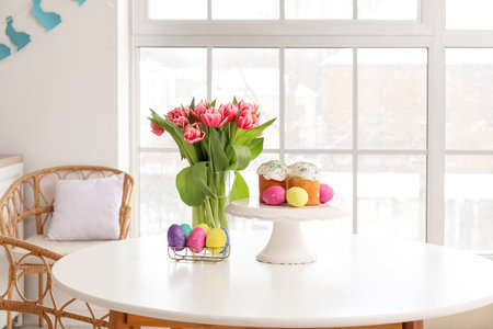 Stand with Easter cakes, eggs and tulips on dining table in kitchen