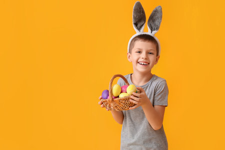 Funny little boy with bunny ears and Easter basket on yellow background