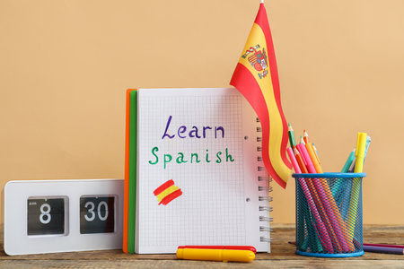 Notebook with text LEARN SPANISH, flag, stationery and clock on table against beige backgroundの写真素材
