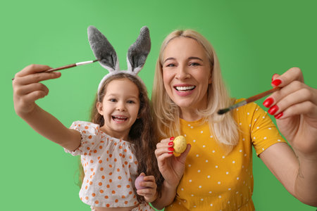 Happy little girl and her grandmother with Easter eggs and brushes on color background
